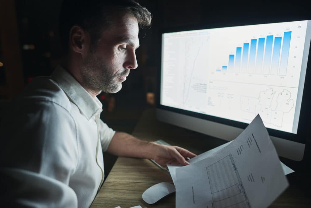 Homem analisando gráficos e documentos em frente ao computador, representando o uso da análise de risco e sustentabilidade na gestão empresarial