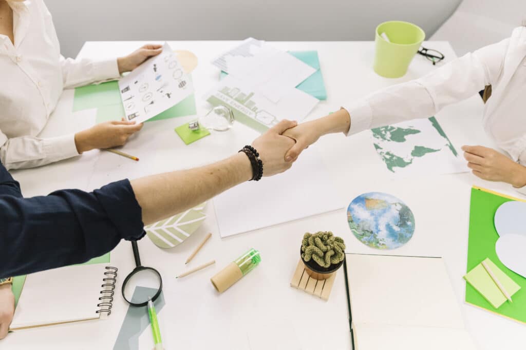 Dois profissionais apertando as mãos em uma mesa com documentos sobre sustentabilidade, incluindo gráficos, mapas e relatórios de sustentabilidade.